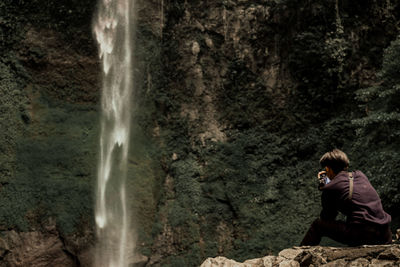 Rear view of man photographing with camera while sitting on rock against waterfall