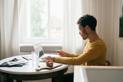 Mid adult man with credit card using laptop while sitting at table in living room