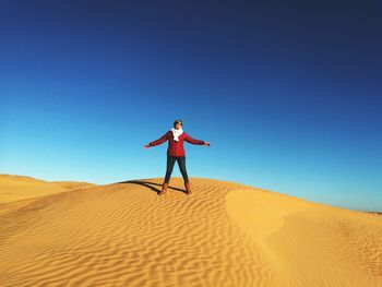 Woman with arms outstretched standing on sand dune in desert against clear blue sky