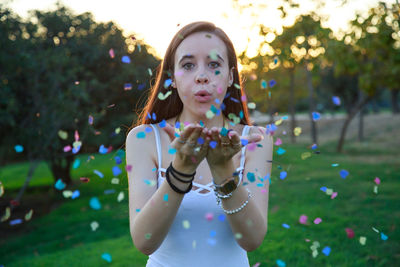Portrait of young woman blowing confetti while standing at park