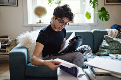 Serious young man doing homework while sitting on sofa with mobile phone and laptop at home