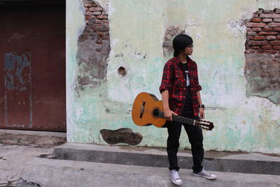 Full length of young woman holding guitar while standing by wall