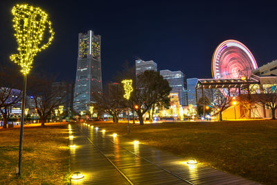 Illuminated ferris wheel by buildings against sky at night