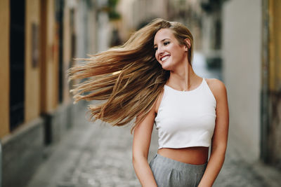 Smiling young woman tossing hair while standing on street amidst buildings