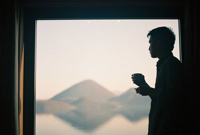 Silhouette man drinking tea while standing against window at home