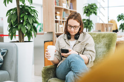 Young teenager girl college or high school student in glasses using mobile phone working at library