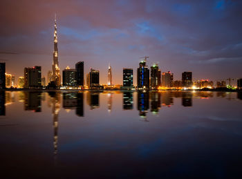 Illuminated buildings by river against sky in city