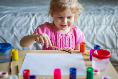 Little smiling girl painting on paper with colorful paints sitting at the table at home