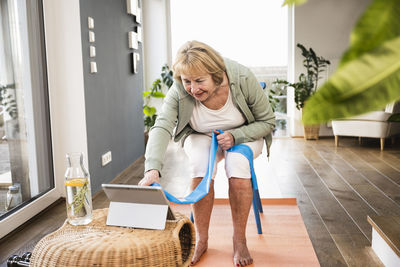 Senior woman with resistance band using tablet pc sitting on chair at home