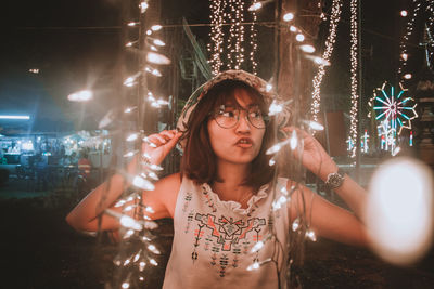 Portrait of young woman standing against illuminated lights at night