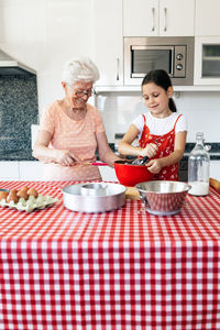 Granddaughter with whisk near grandmother in eyewear cooking dough at table in house