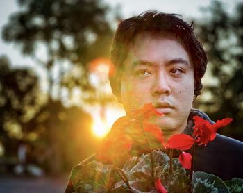 Young asian man holding red cyclamen potted plant against trees and setting sun.