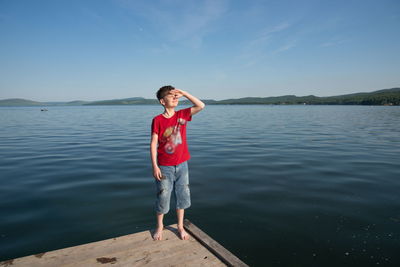 A boy stands on a wooden pier and looks at the sun from under his arm, against lake on a sunny day.