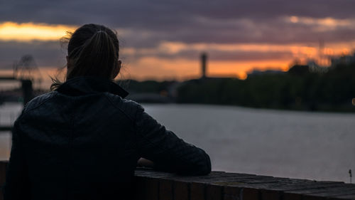 Rear view of woman leaning in railing by river against cloudy sky during sunset