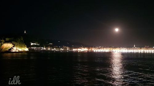 Illuminated city by sea against sky at night