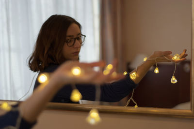 Reflection of woman holding illuminated string lights in mirror