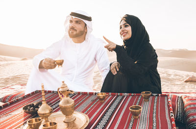 Smiling woman gesturing by man with tea sitting on carpet at desert