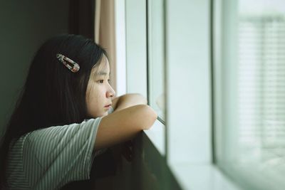 Side view of thoughtful teenage girl looking through window at home