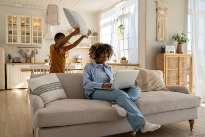 Calm smiling african american mother freelancer sits on couch working on laptop with child son