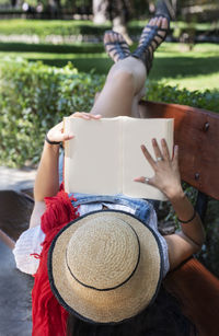 Woman holding book while lying on bench in park