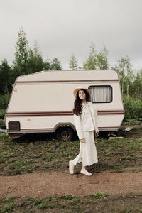 Side view of young woman standing by car against sky