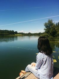 Rear view of woman sitting on pier over lake against clear blue sky