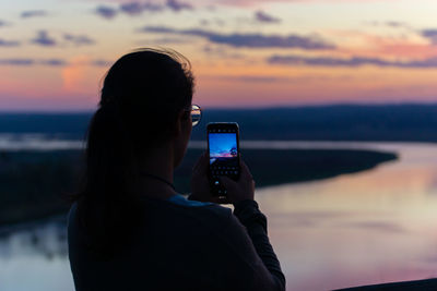 Rear view of man photographing sea against sky during sunset