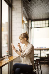 Woman applying lipstick while sitting on stool in bar