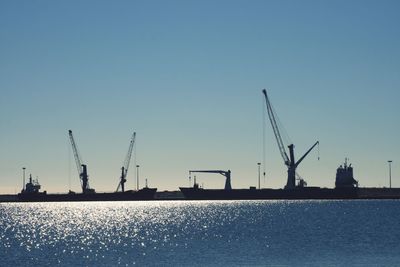 Silhouette cranes at commercial dock against clear blue sky