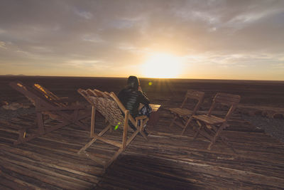 Rear view of person sitting on wooden chair against sky at sunset