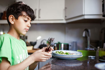 Side view of boy holding forks while standing at kitchen counter