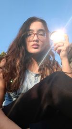 Portrait of young woman holding eyeglasses against sky