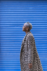Side view of cheerful african american female with short hair in coat looking away with smile while standing on blue background in city