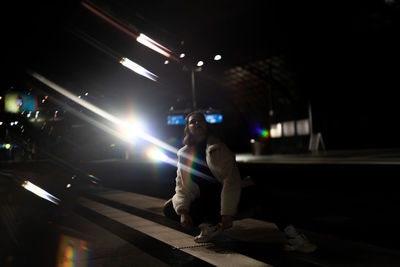 Woman crouching on street against illuminated lights at night