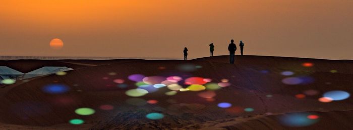 Silhouette people standing on sand by illuminated lights against sky during sunset