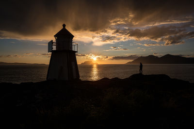 Silhouette man standing by lighthouse at shore against cloudy sky during sunset