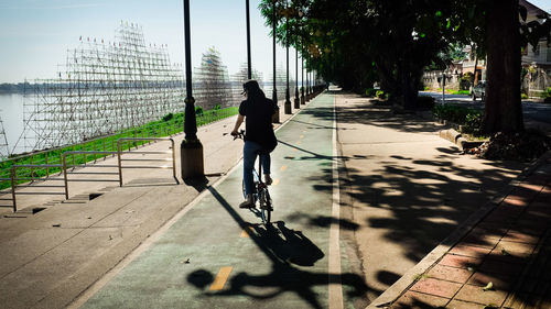 Rear view full length of woman riding bicycle on footpath during sunny day