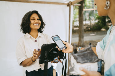 Happy female owner communicating with customer doing mobile payment at flea market