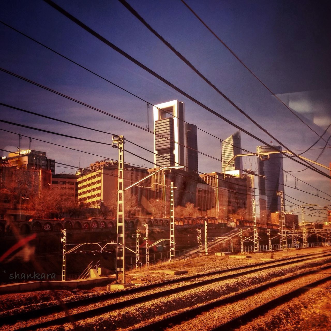 railroad track, power line, building exterior, electricity pylon, built structure, architecture, rail transportation, power supply, sky, cable, electricity, city, connection, transportation, public transportation, no people, railway track, cityscape, fuel and power generation, outdoors