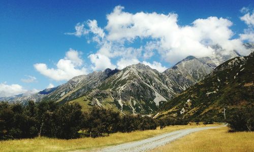 Country road leading towards mountains against cloudy sky