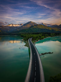 Road leading towards mountains by lake against sky during sunset