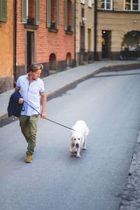 Full length of senior man walking with dog on road by buildings in city