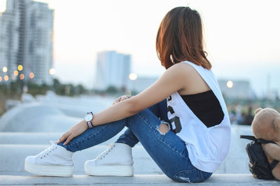 Side view of woman sitting on railing in city against clear sky during sunset