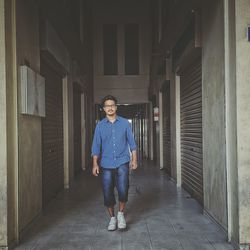 Portrait of young man walking on corridor amidst closed shutters