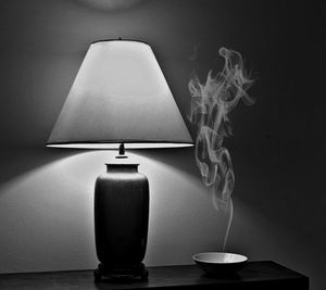 Smoke from bowl by illuminated lamp shade on table against wall at home