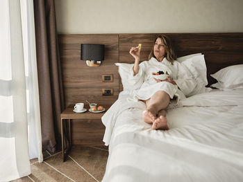 Senior woman eating fresh fruits in breakfast while relaxing on bed and looking away at hotel room