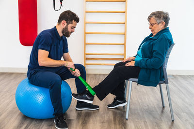 Side view of male personal coach sitting on exercise blue ball while using green elastic band on ankle of old woman in gym