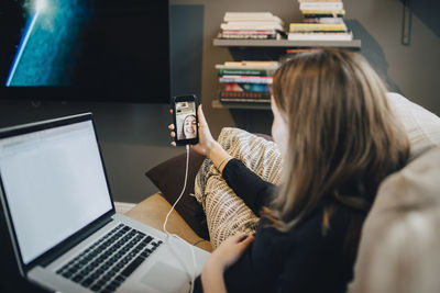 Girl taking selfie through mobile phone while using laptop on sofa