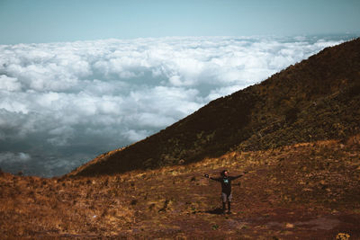 High angle view of young man with arms outstretched standing on mountain against sky