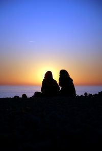 Silhouette couple sitting on beach against sky during sunset
